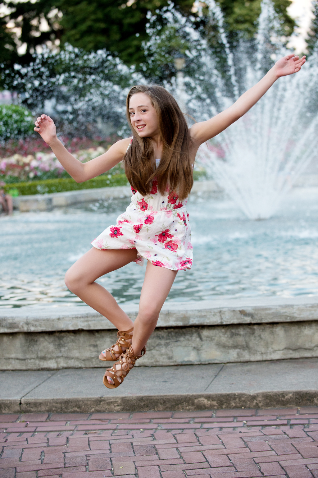 a young girl jumps in the air in front of a water fountain and clicks her heels together