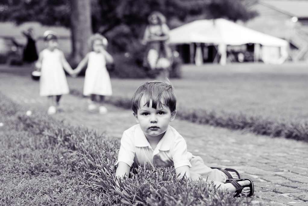 ring bearer lays in the bushes as flower girls walk down aisle behind him