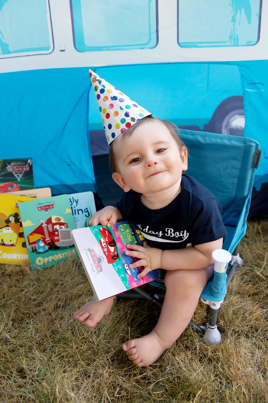a baby boy sits in a camp chair with a polka dot birthday hat on