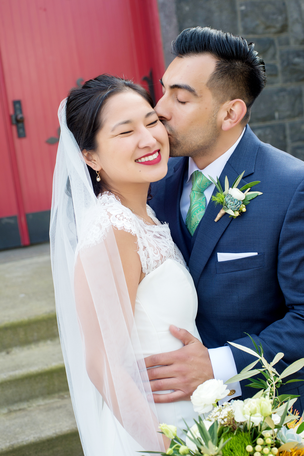 groom kisses brides cheek in front of a red door