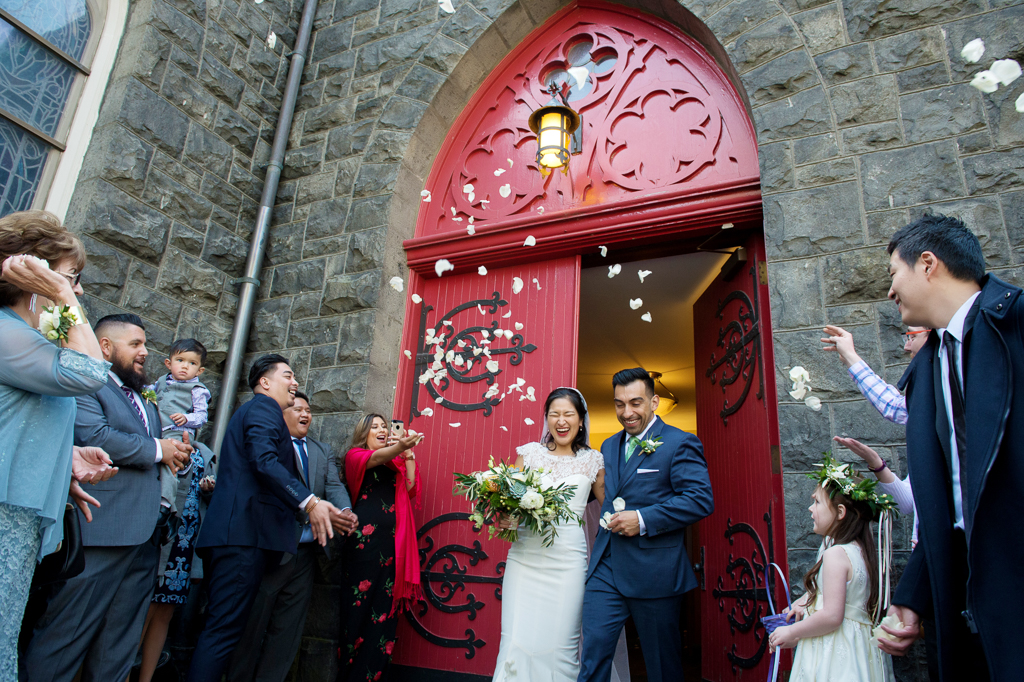 wedding guests throw white flower petals at the newly married bride and groom as they exit the large red doors of trinity episcopal church