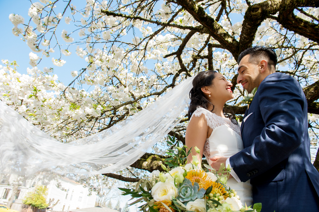 bride and groom look at each other underneath a white cherry blossom tree as veil flies in wind