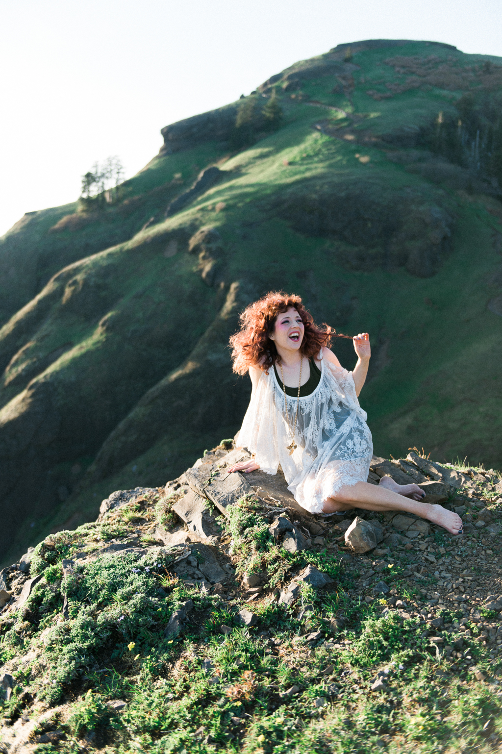 a woman with wild red hair sits on a rock on the top of saddle mountain wearing a white lace dress