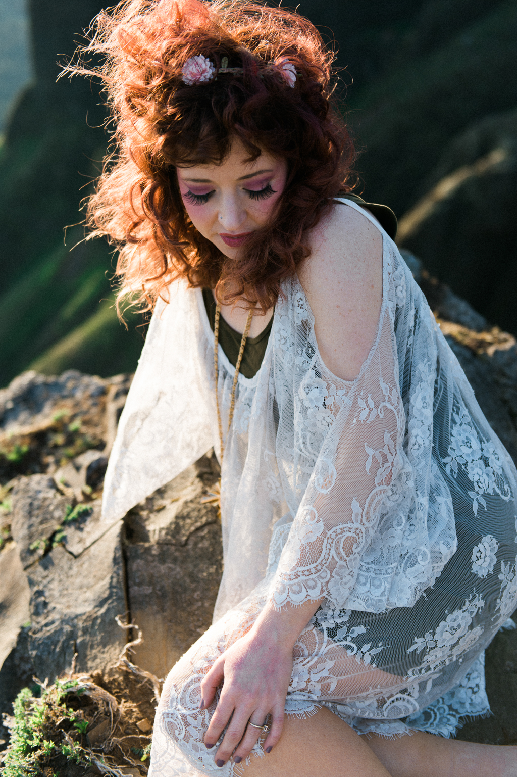 a woman with wild red hair sits on a rock on the top of saddle mountain wearing a white lace dress