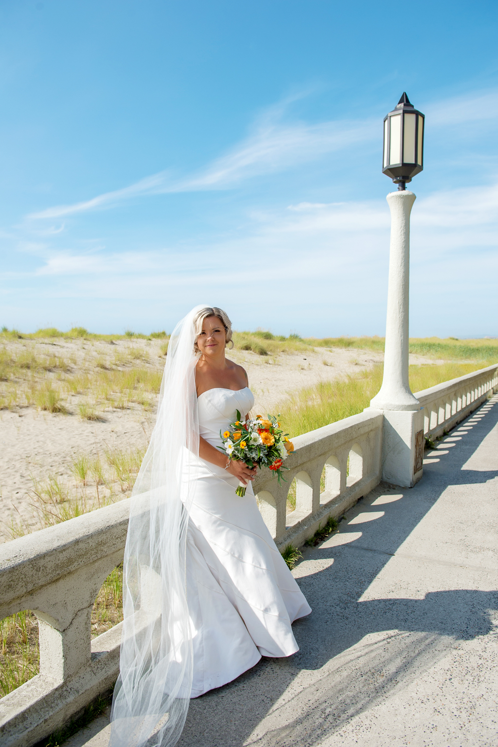 bride stands on boardwalk holding yellow and orange bouquet