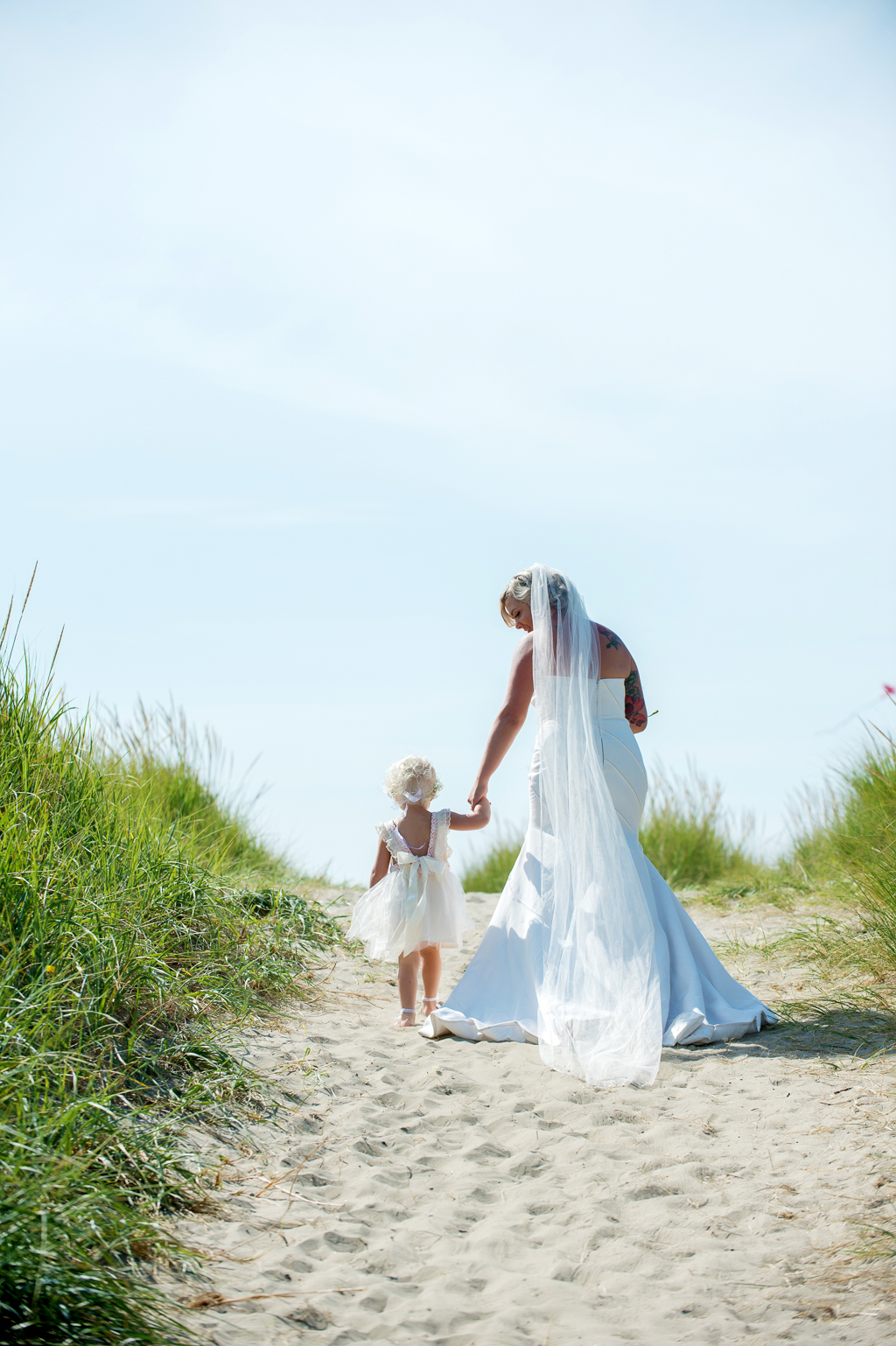 bride holds flower girl's hand as they walk over a sand dune on the beach