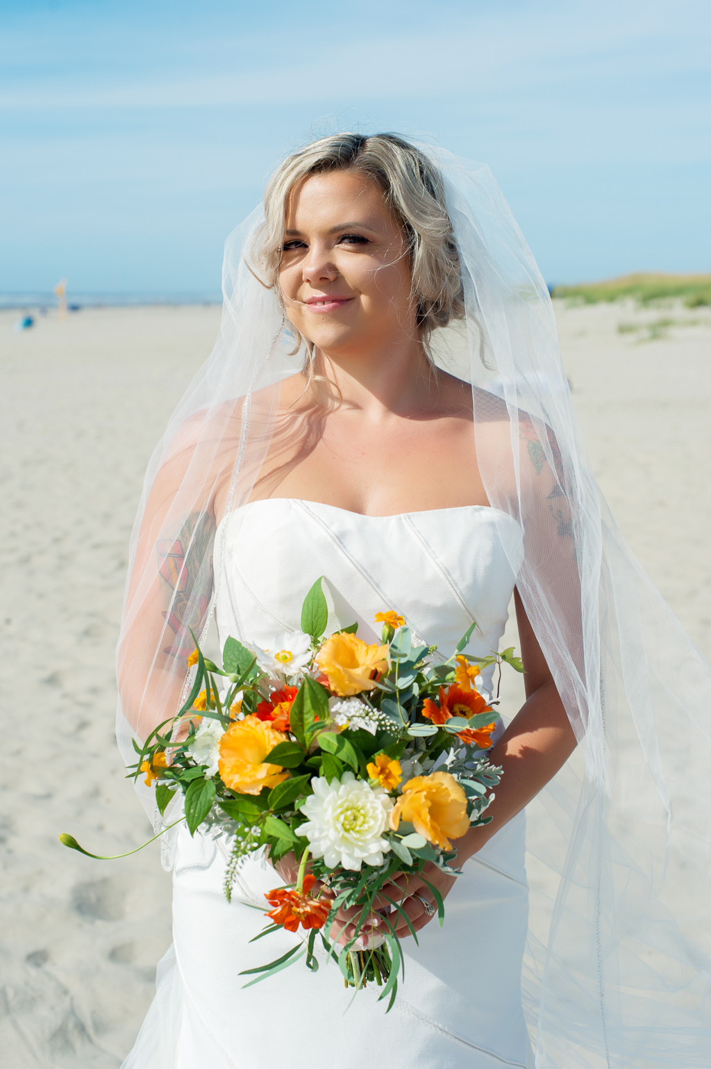 bride stands on beach holding yellow and orange bouquet