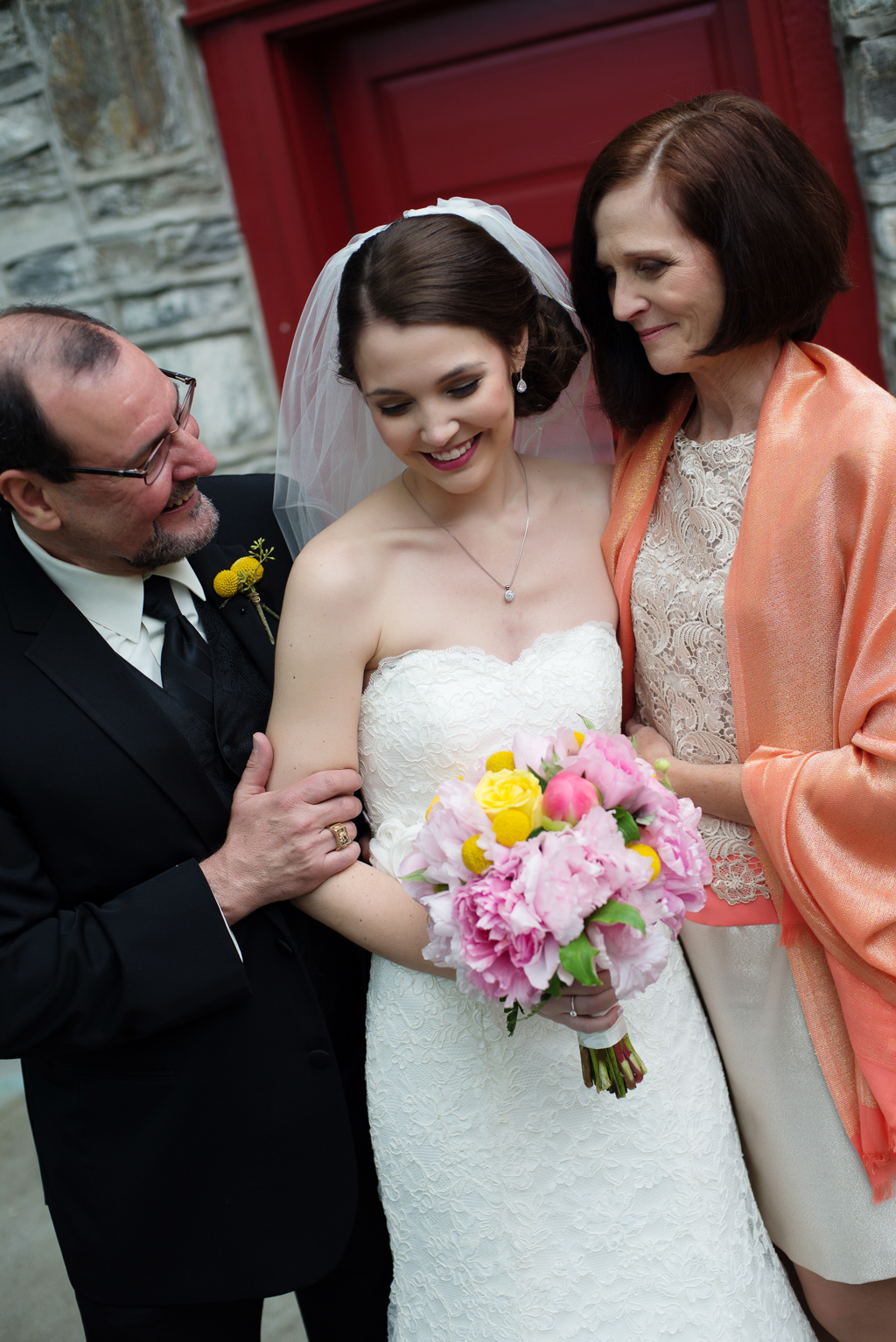 a mother and father hug lovingly around their daughter on her wedding day as she holds a pink and yellow bouquet
