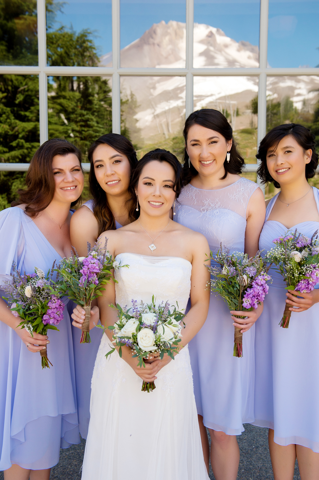 bridesmaids in purple dresses hug around bride with reflection of mt hood in the window behind them
