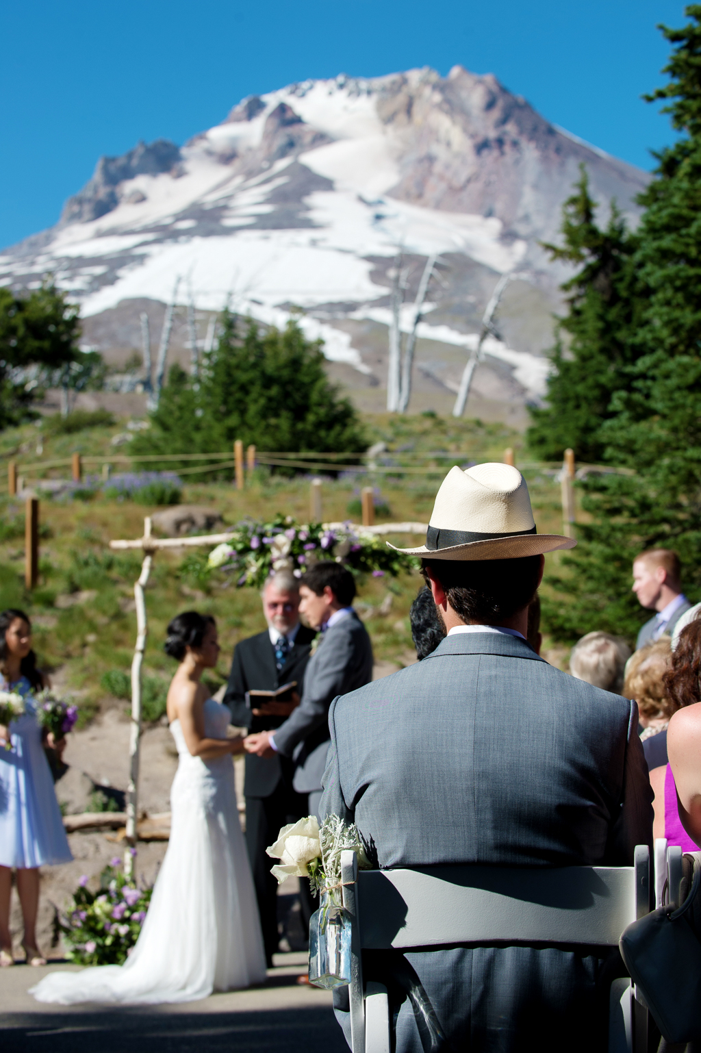 a man in a fancy hat looks onto a wedding ceremony behind timberline lodge with mount hood towering above