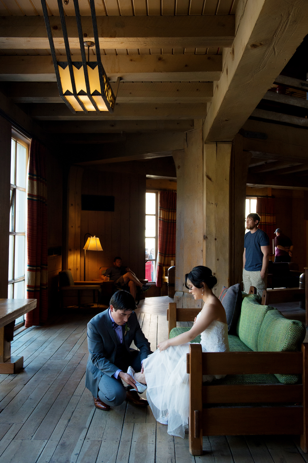 groom helps fit bride's shoes in timberline lodge as she sits on a green wooden couch