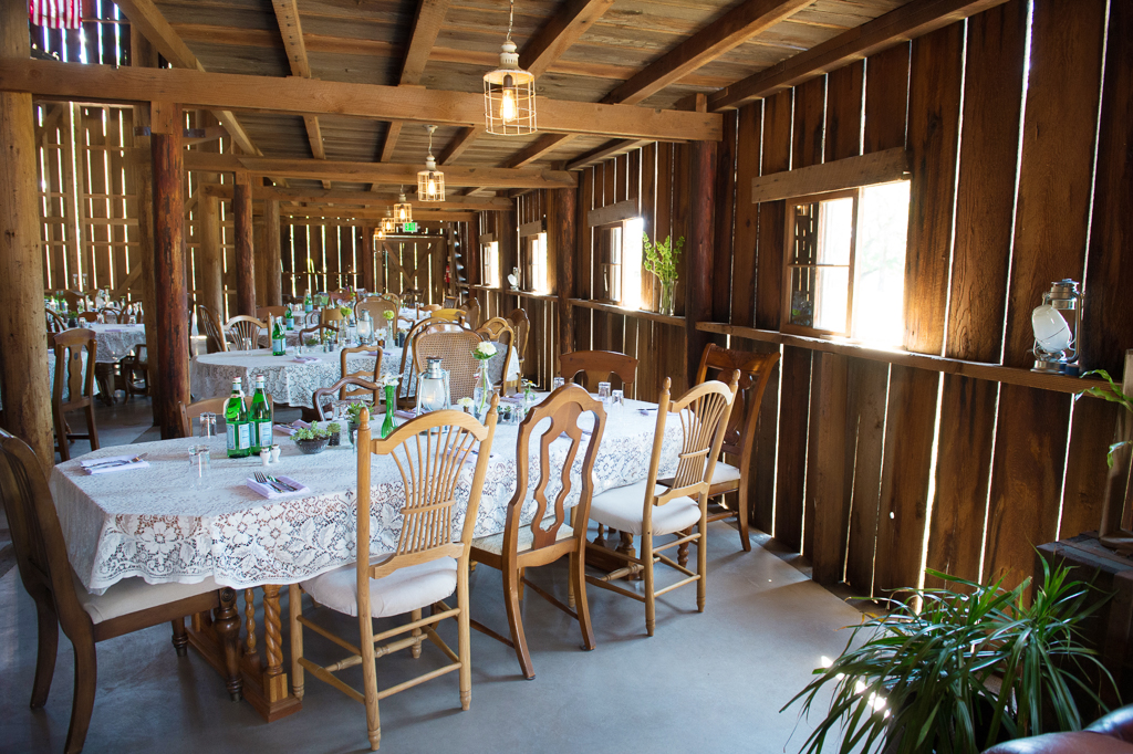 tables covered in lace and filled with small glass bottles trinkets and flowers fill a giant wooden barn for a wedding reception