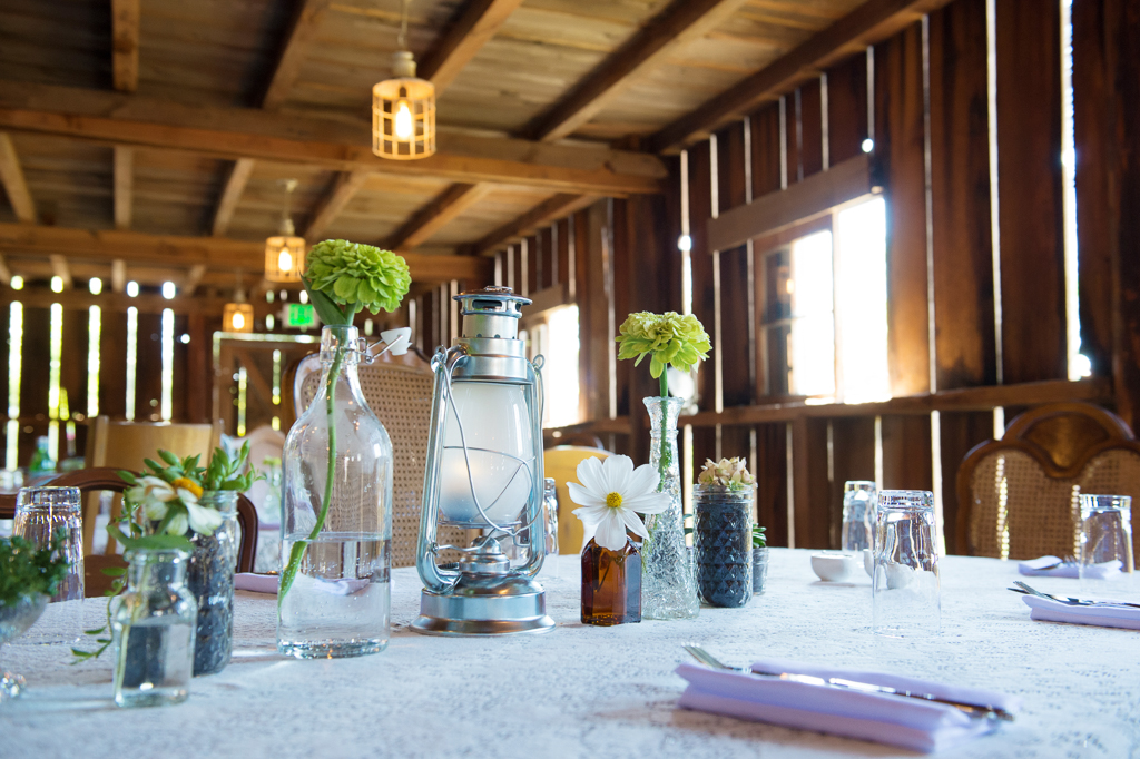 tables covered in lace and filled with small glass bottles trinkets and flowers fill a giant wooden barn for a wedding reception