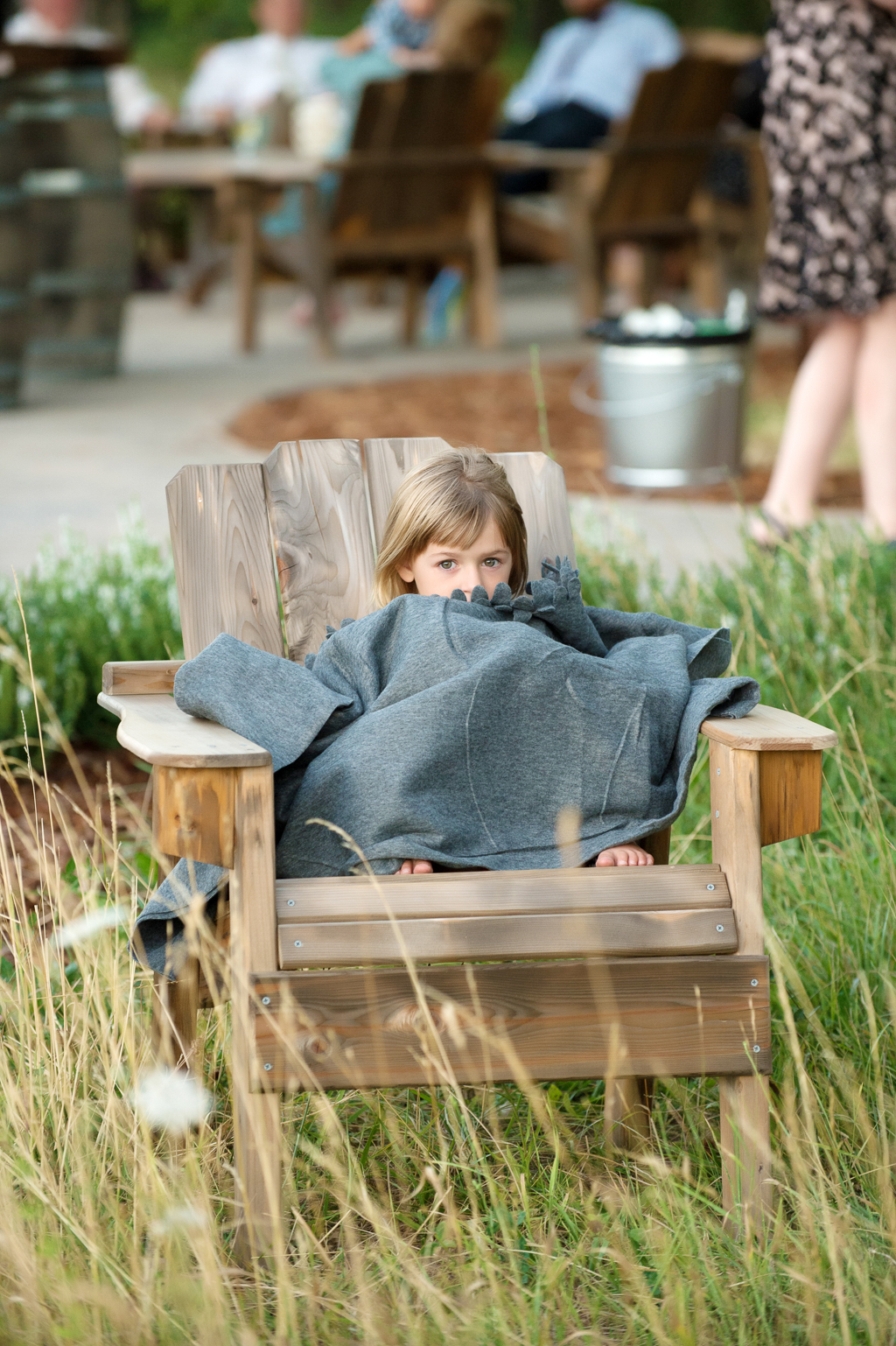 a little girl sits in a wooden adirondack chair outside wrapped in a blanket