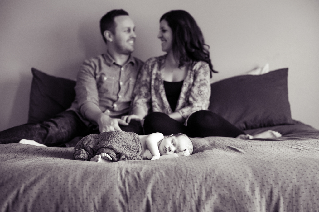 a newborn baby lays on the foot of the bed while mom and dad hug together