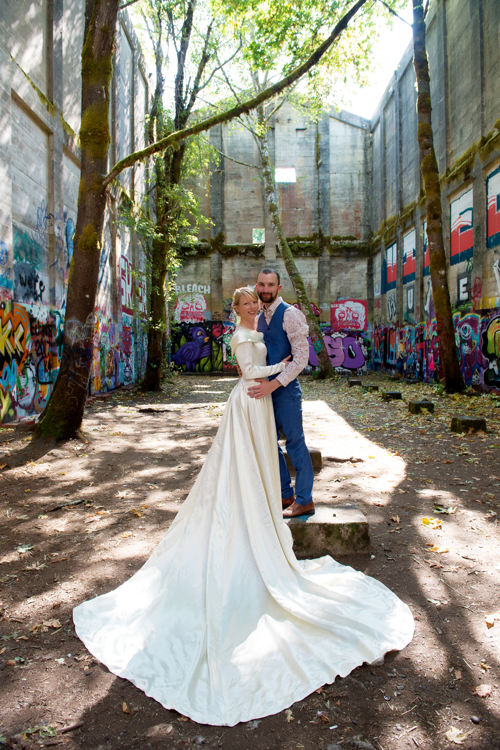 bride in a long sleeved wedding dress with a huge train and groom stand in the middle of an old mill in vernonia that is covered in graffiti