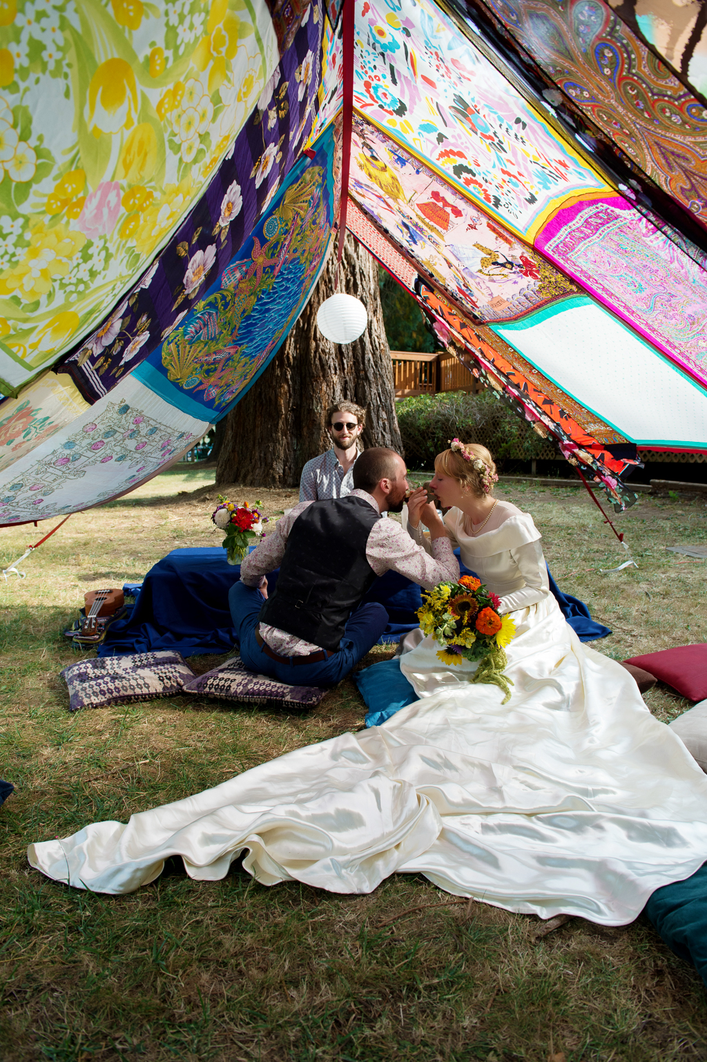 a bride and groom sit on pillows on the ground sipping tea under a makeshift tent made of colorful scarves