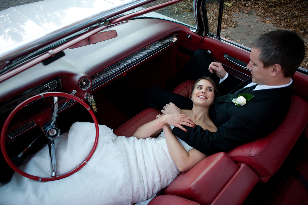 bride lays in groom's lap across front seat of vintage cadillac convertible with red leather interior
