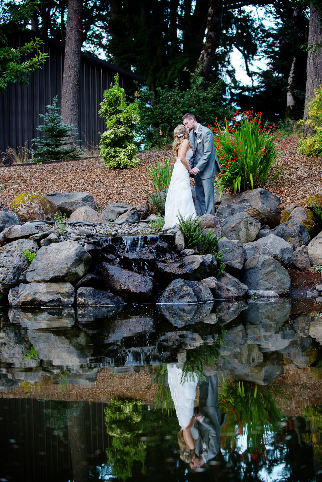 a bride and groom stand on a pile of stones next to a pond with their reflection in the water