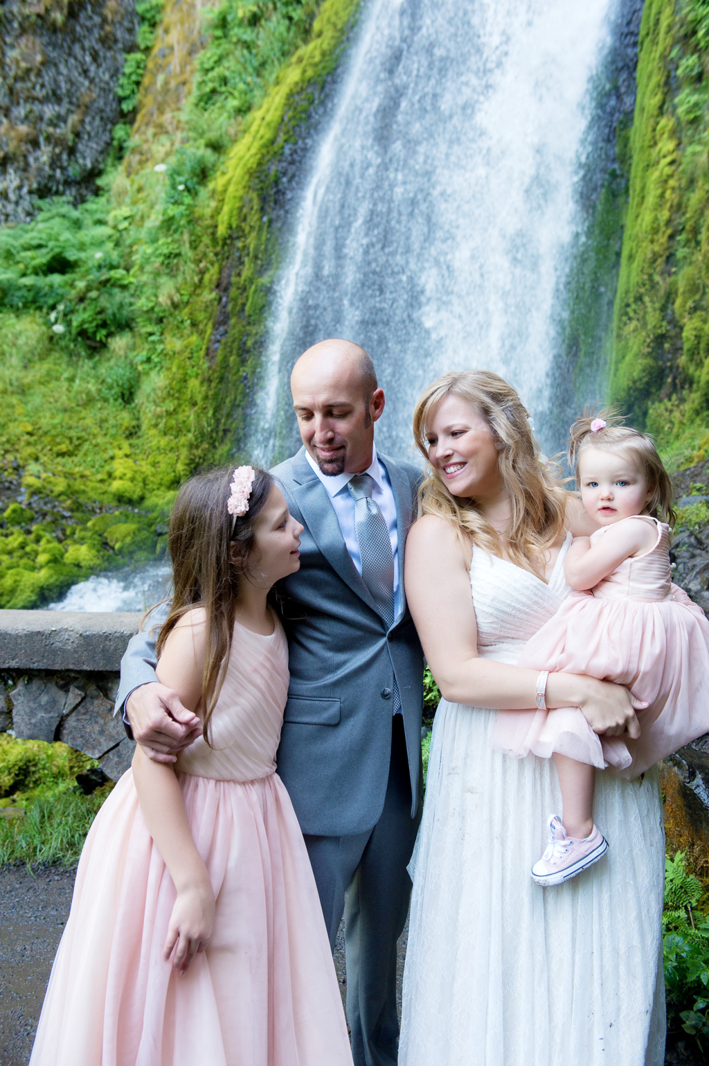 a bride and groom hug around their daughters wearing pink dresses in front of wahkeena falls