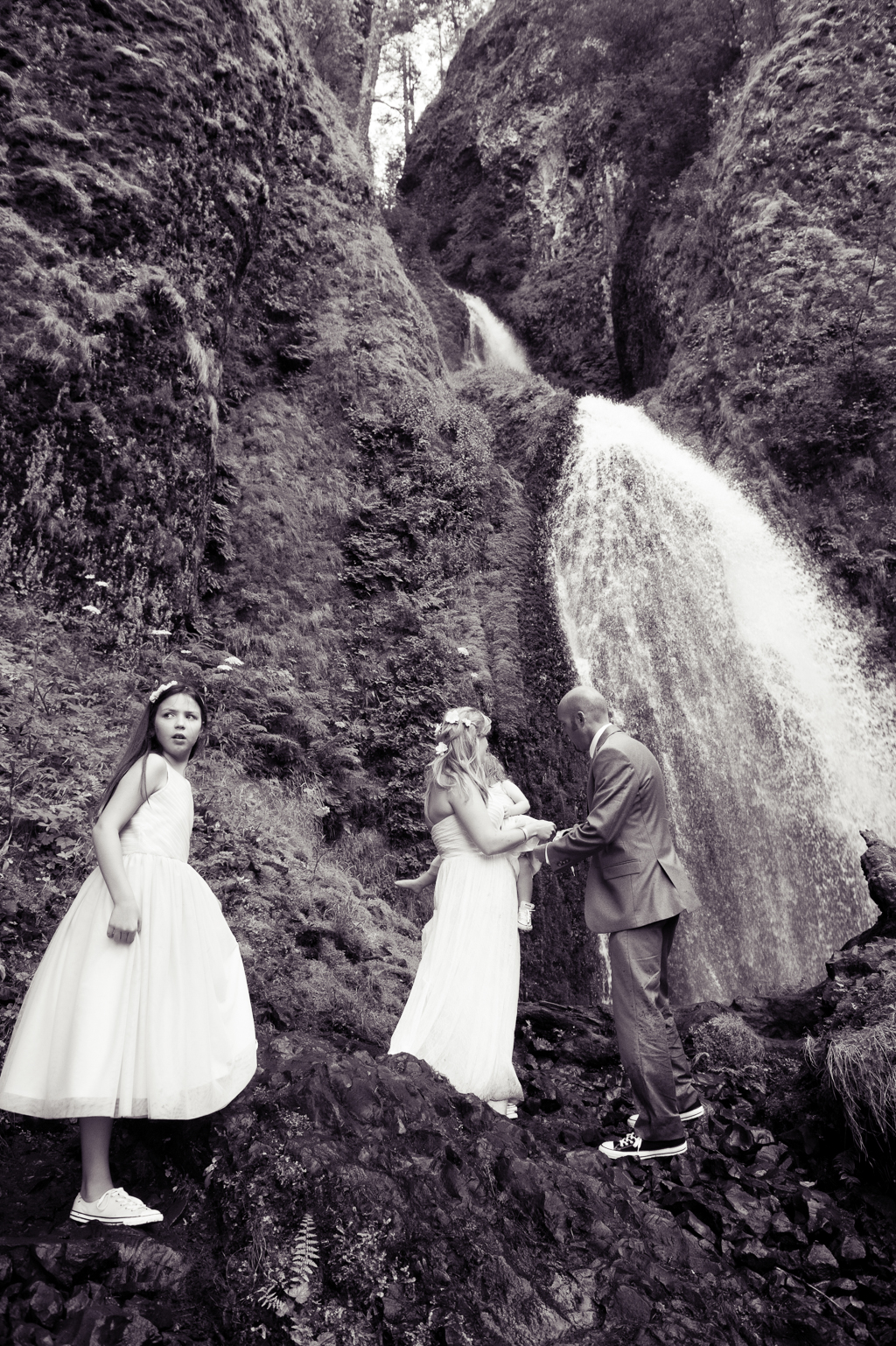 a groom helps a bride climb rocks at wahkeena falls as the flower girl looks over her shoulder