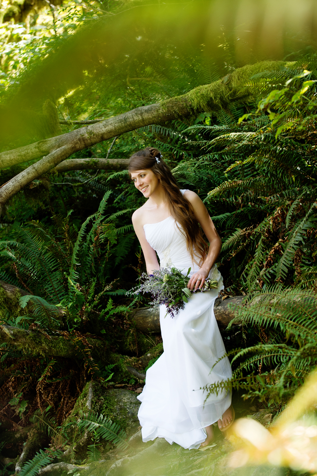 a bride holds a purple and lavender bouquet and sits on a moss covered tree log in the forest