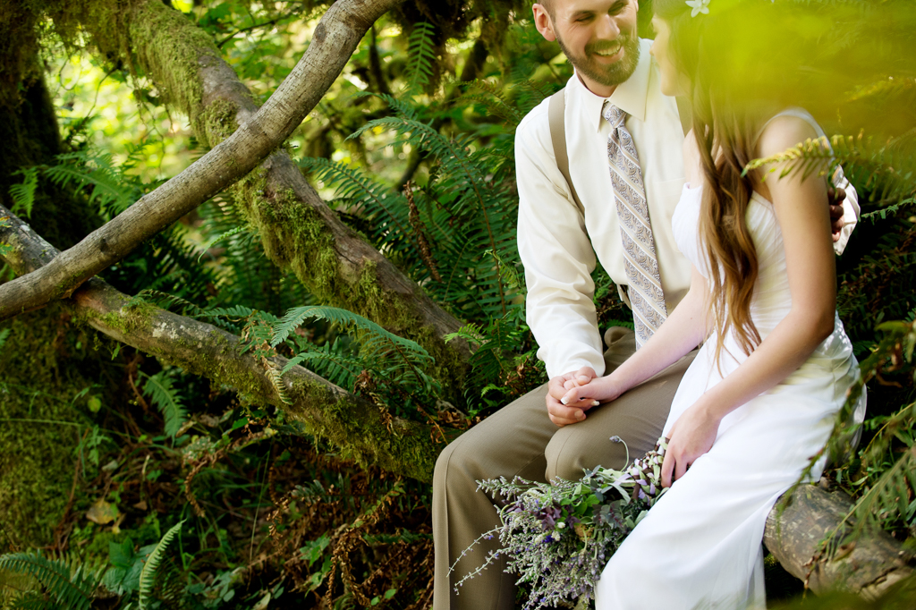 a bride holds a purple and lavender bouquet and sits on a moss covered tree log in the forest with her groom
