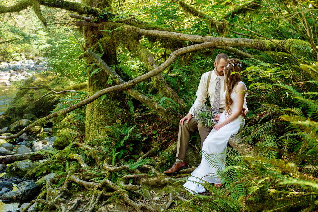 a bride holds a purple and lavender bouquet and sits on a moss covered tree log in the forest kissing her groom