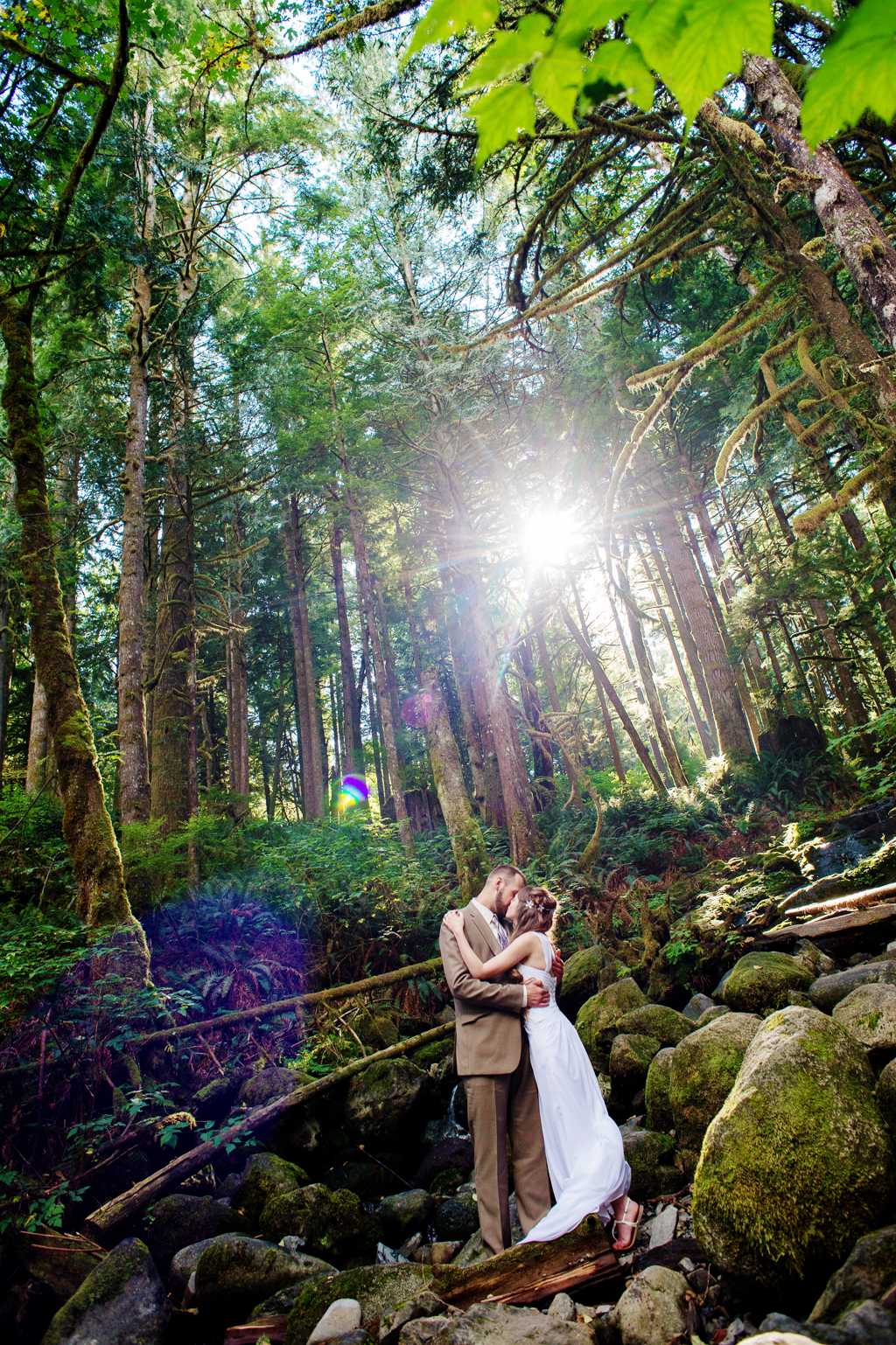 a bride and groom embrace and kiss in a magical mossy forest as the sunlight shines through