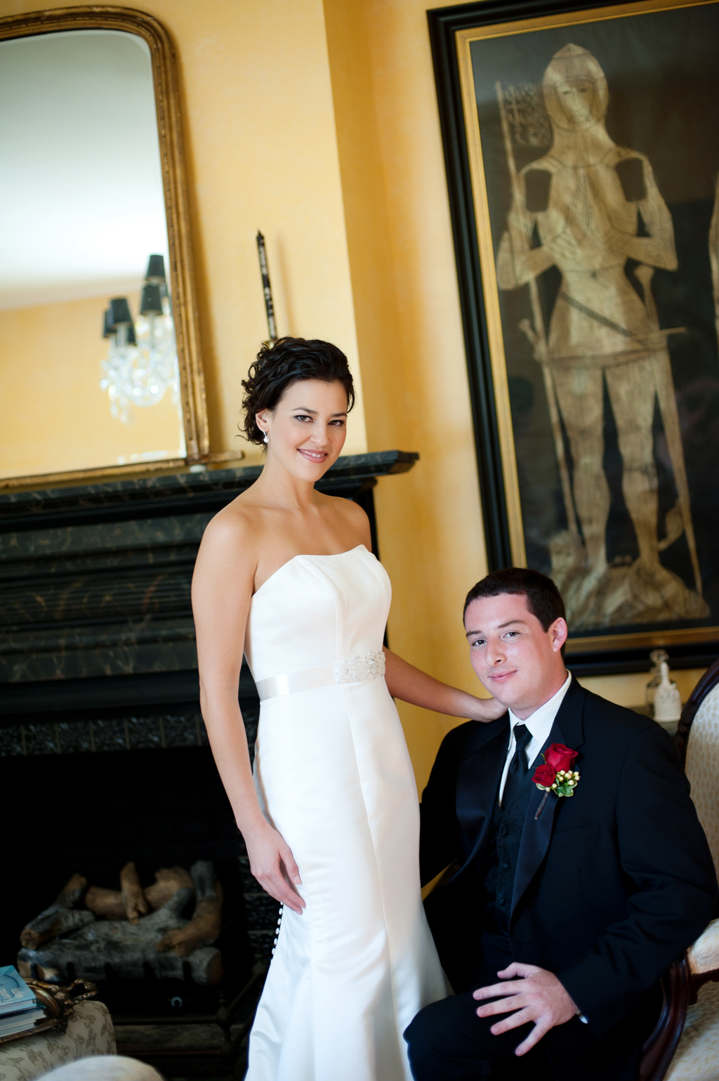 a groom sits in an antique chair and a bride stands over his shoulder in a stately yellow room with very old art on the wall