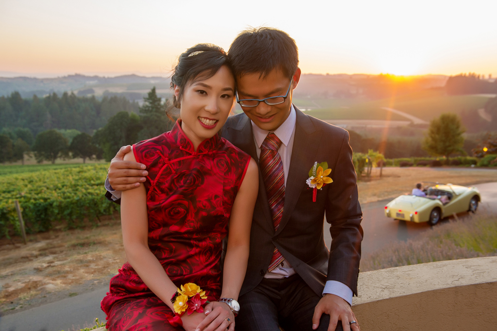 bride in red wedding dress and groom overlook willamette valley vineyards at sunset as a vintage yellow convertible drives by