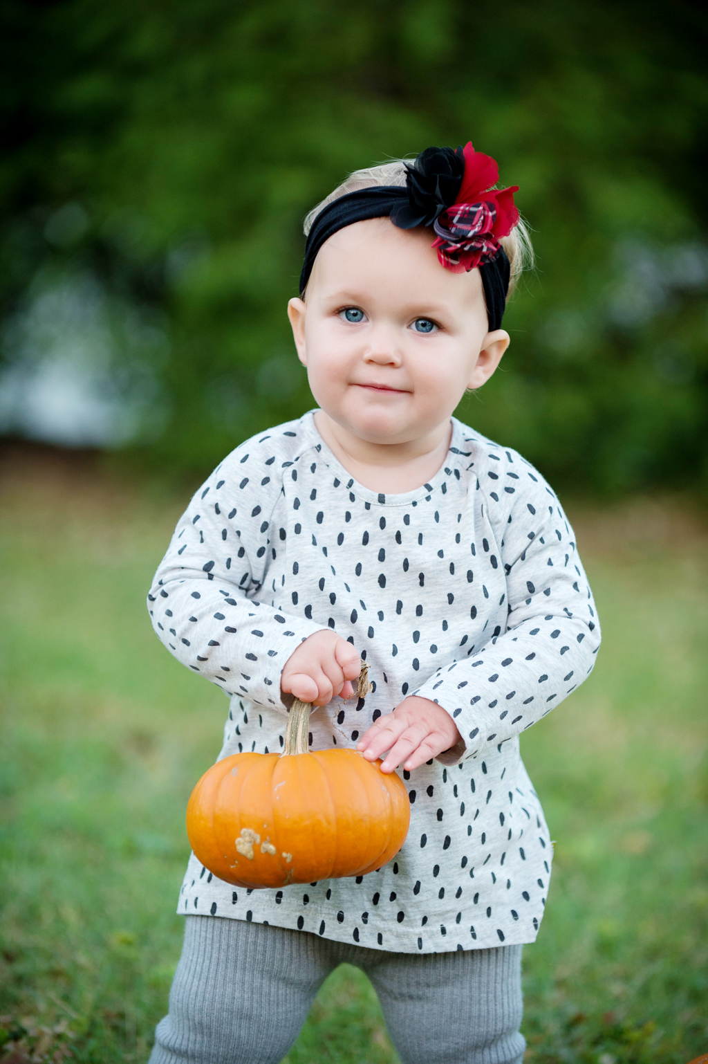 a baby girl in a black and white polka dotted shirt holds a pumpkin