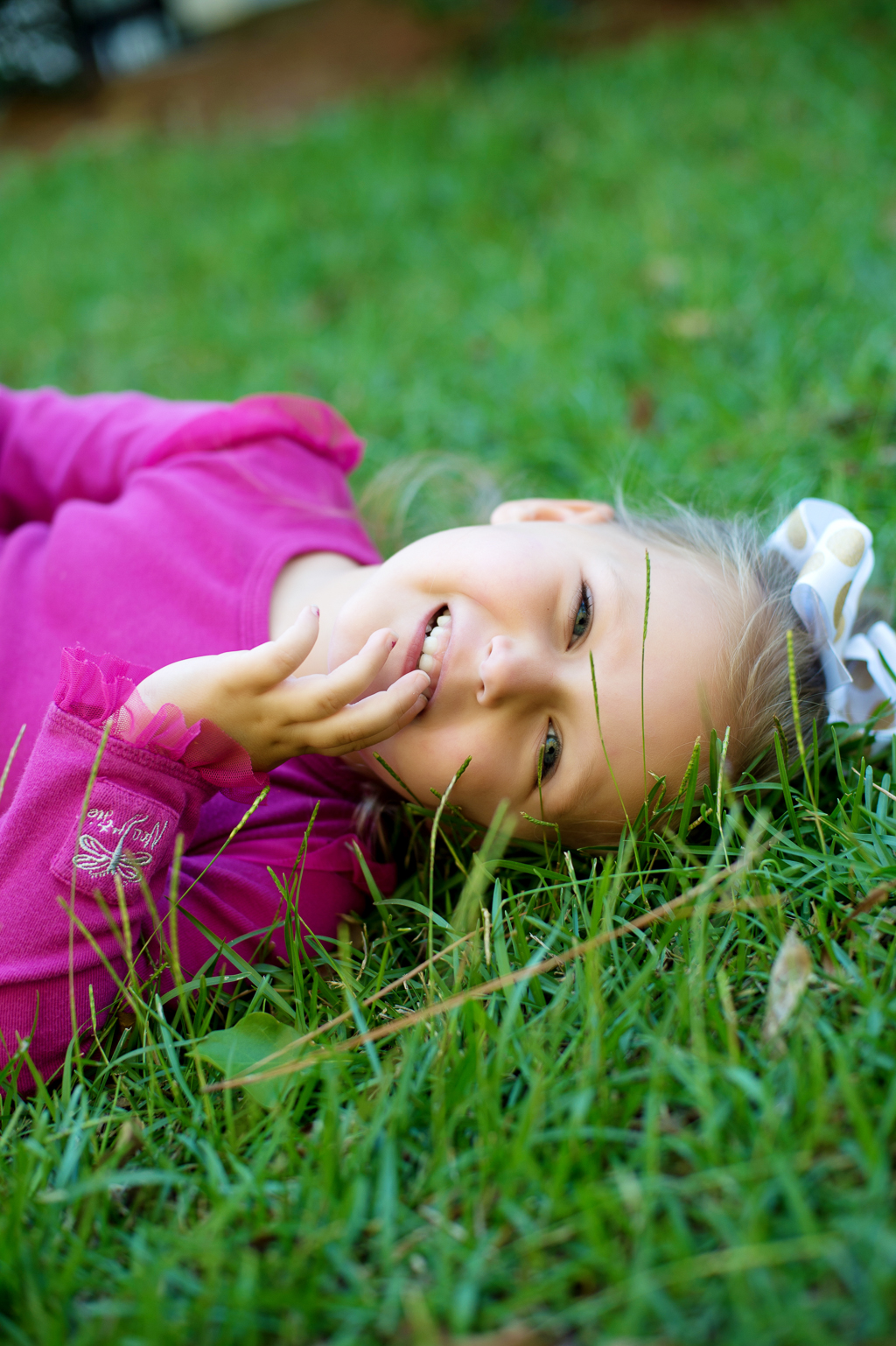 a young girl in hot pink lays in the grass and smiles with her hand to her face