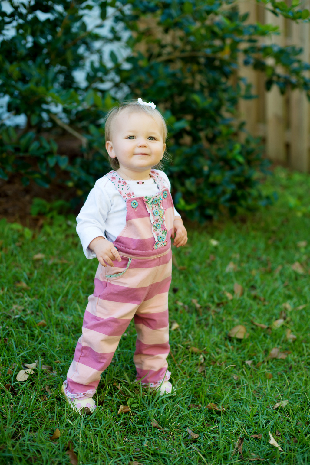 a baby girl in a pink striped overalls stands smiling in the grass
