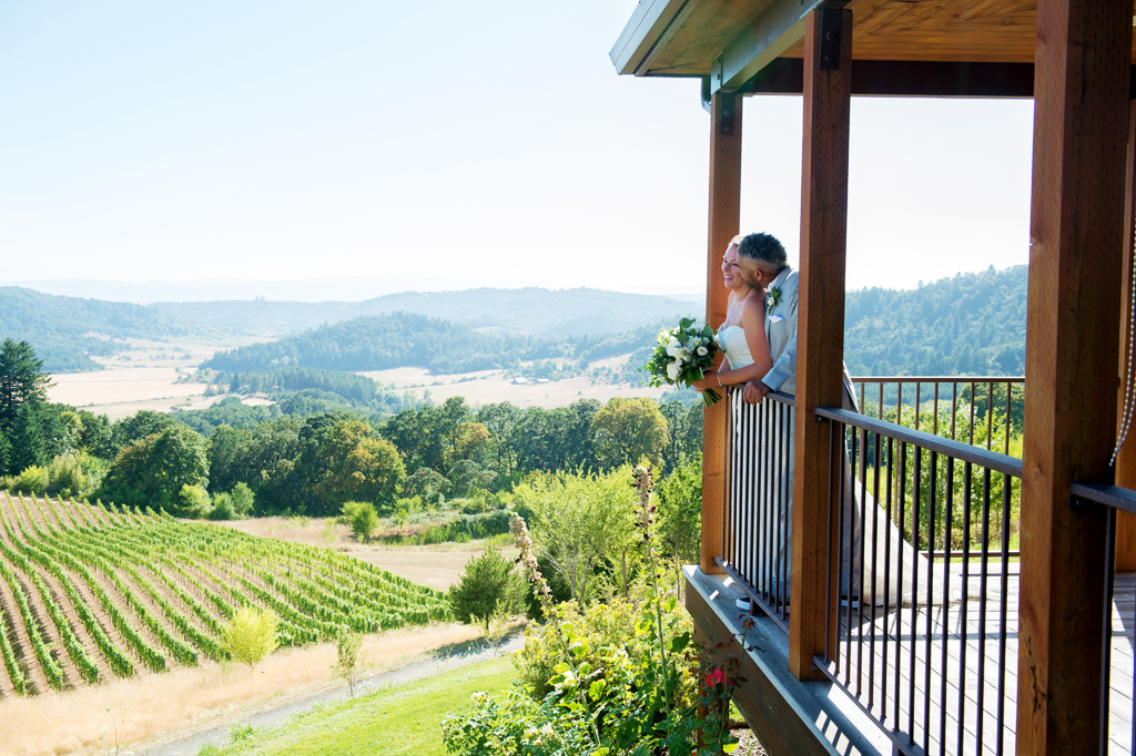 a bride and groom stand over the porch railing looking off at youngberg hill vineyard