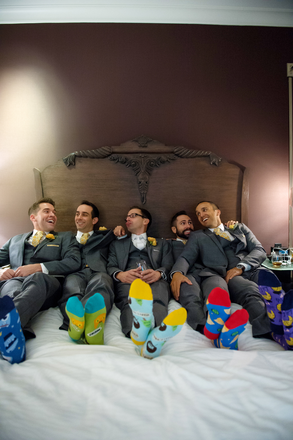 a groom and groomsmen lay across a hotel bed showing off their happy feet socks