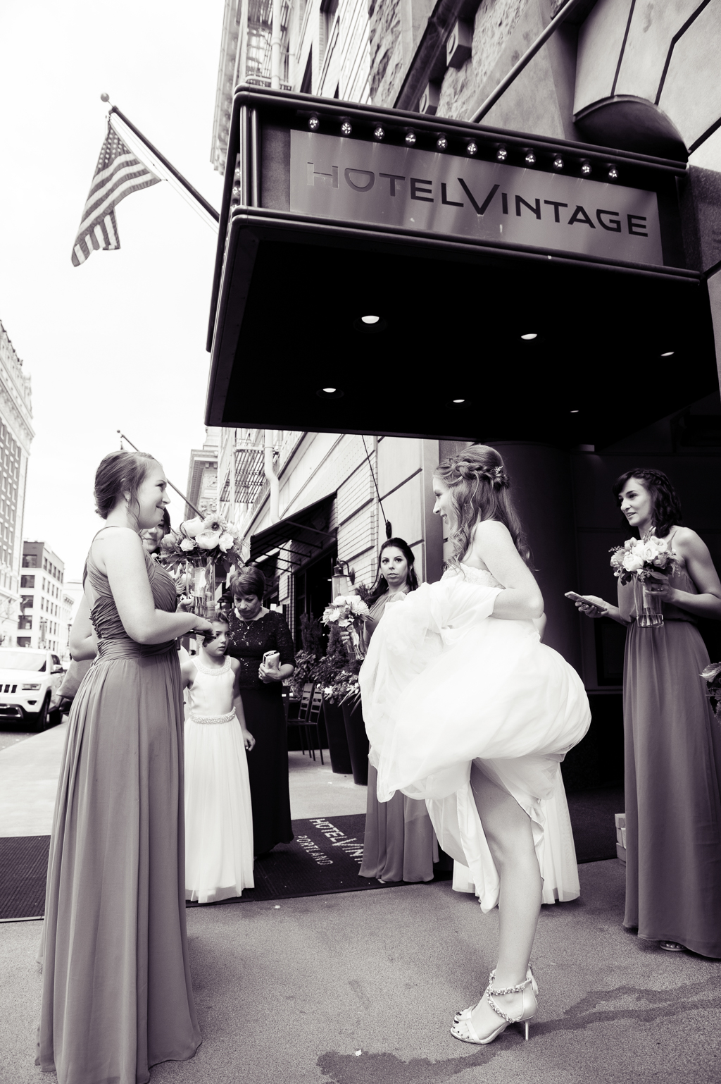 bride stands outside front entrance of hotel vintage holding her wedding dress up while her bridesmaids wait for taxi