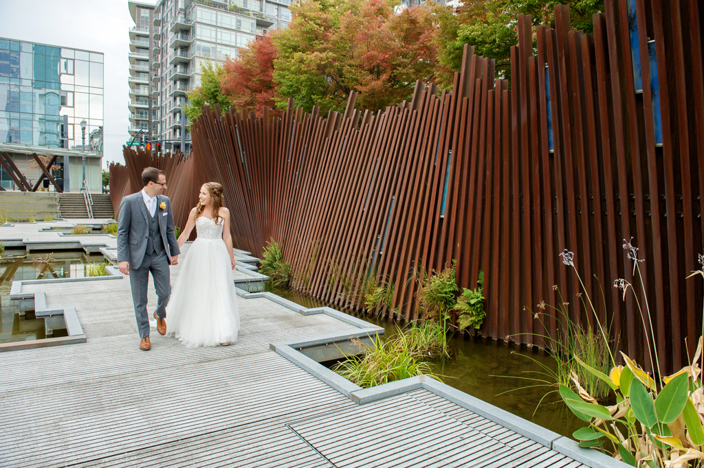bride and groom hold hands and walk along boardwalk in front of rusty fence sculpture wall at tanner springs park