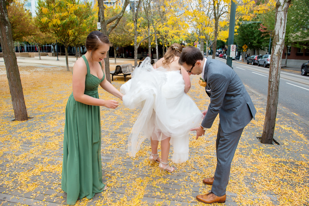 bridesmaid in a green dress and groom help bride pick yellow leaves out of her wedding dress train