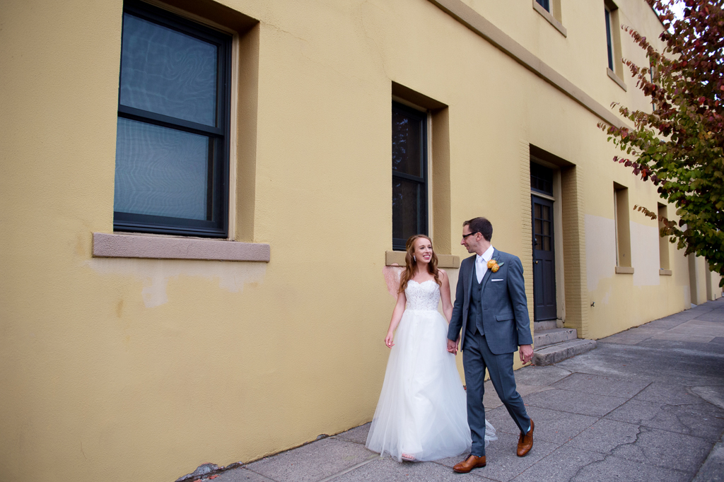 a bride and groom walk hand in hand in front of a mustard yellow wall