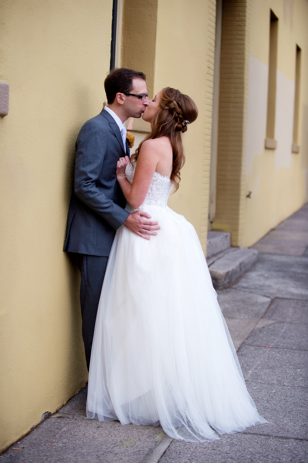 a groom leans against a yellow wall and kisses his bride