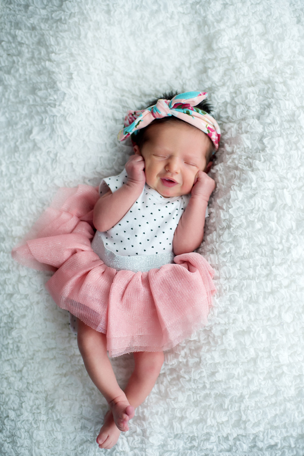 A newborn girl in a pink tutu and polkadot top smiles with her hands on her cheeks