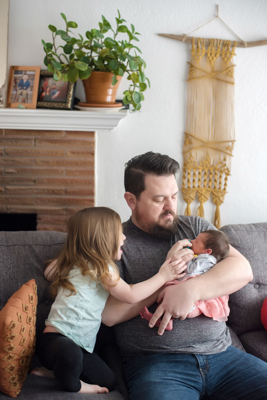 a dad holds a newborn girl as her big sister looks over his shoulder at her sitting on their living room couch in their home