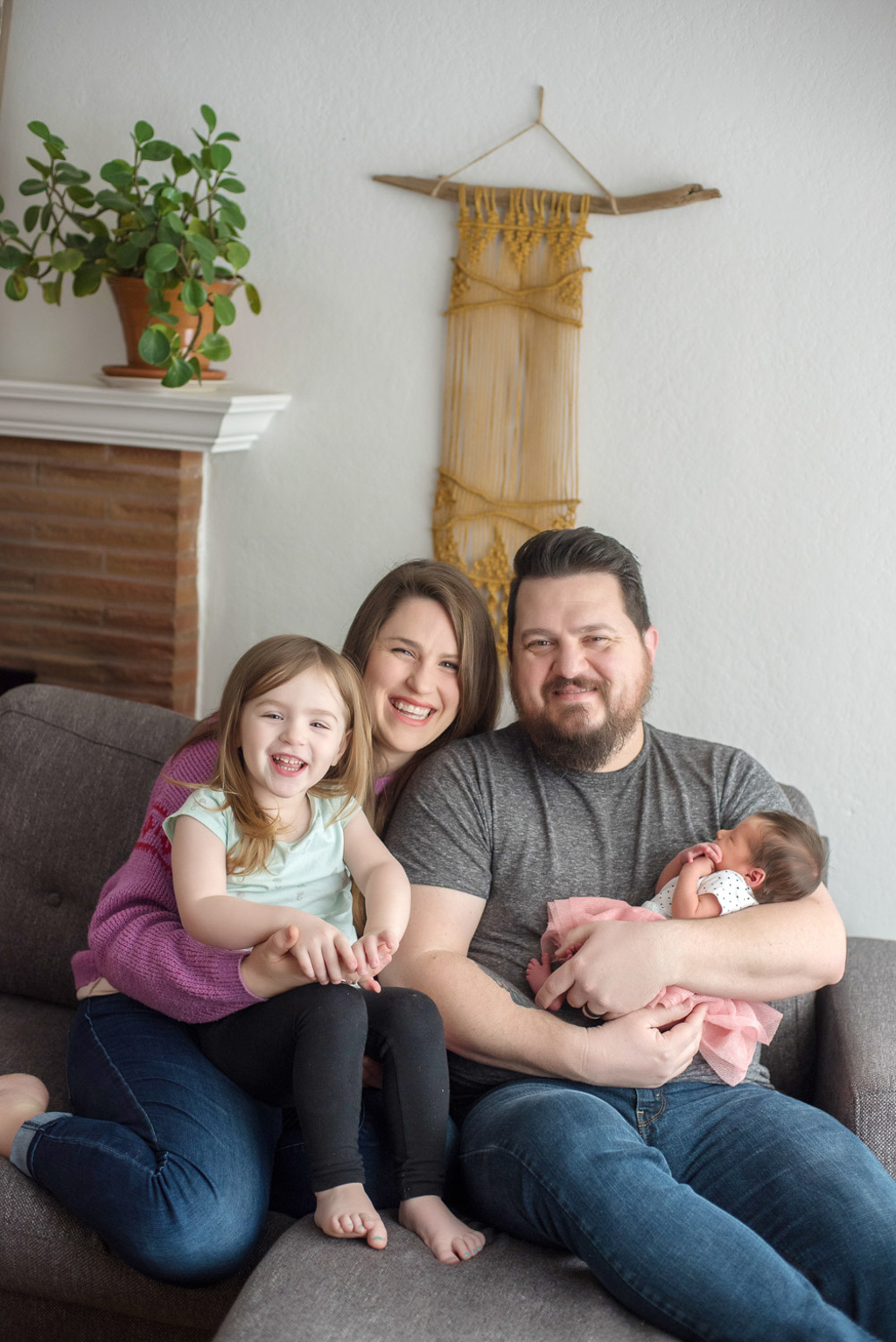 a mom and dad sit on their living room couch in their home holding their newborn daughter and 2 year old daughter
