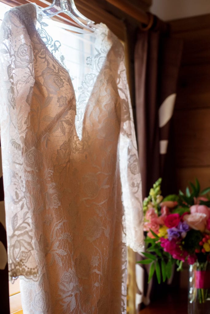 a long sleeved lace wedding dress hangs from a window with a colorful bouquet in the background
