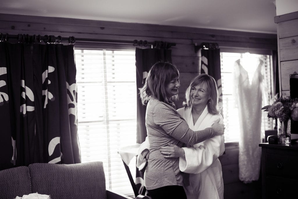 a woman happily embraces another woman who is getting ready for her wedding day.  a bridal gown hangs in the window in the background
