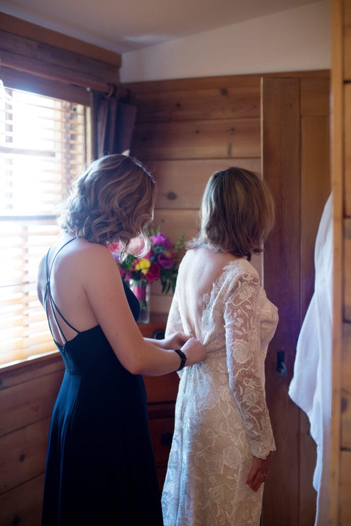 a young woman helps her mother button up her wedding dress 