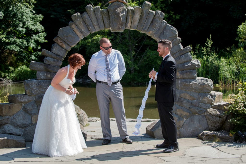 the groom pulls out a very long piece of paper with his vows in front of the stone arch during their wedding ceremony 