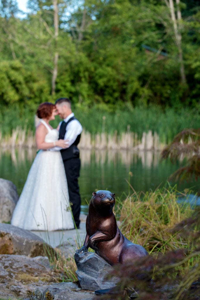 a bronze sea otter sculpture is in the foreground with a bride and groom embracing by the pond in the background 