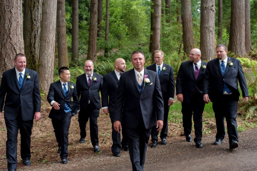 groomsmen in navy and purple walk and laugh underneath large trees
